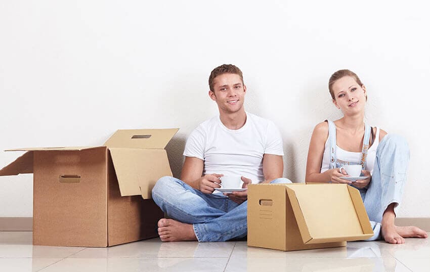 The image depicts a Hire Man and Van removal van parked on a residential driveway in Whitburn, with its side door open revealing a collection of various discarded household items. Visible in the cargo area are a stack of cardboard boxes, some partially crushed and layered, alongside a few black plastic refuse bags filled with waste. Leaning against the inside wall of the van are a couple of wooden pallets and a rolled-up carpet with a textured, fabric finish. The driveway surface appears to be made of concrete, bordered by a garden area with sparse vegetation visible in the background. The scene includes a portion of the front of a terraced house with a brick façade, typical of suburban properties in the EH47 postcode area. Overcast daylight provides diffuse lighting, with soft shadows cast across the scene. The overall composition highlights the process of rubbish removal, aligning with services offered by Hire Man and Van for local household clearance needs in Whitburn and nearby locations.
